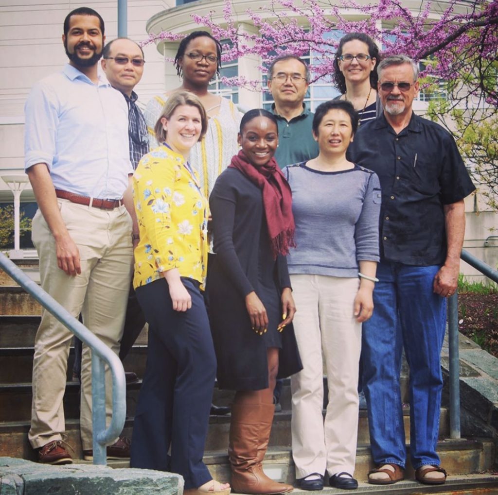 Photo of Kizzmekia Corbett and team: A very diverse group of UMBC alumni and research faculty smile together for a photo in front of a tree with purple flowers.