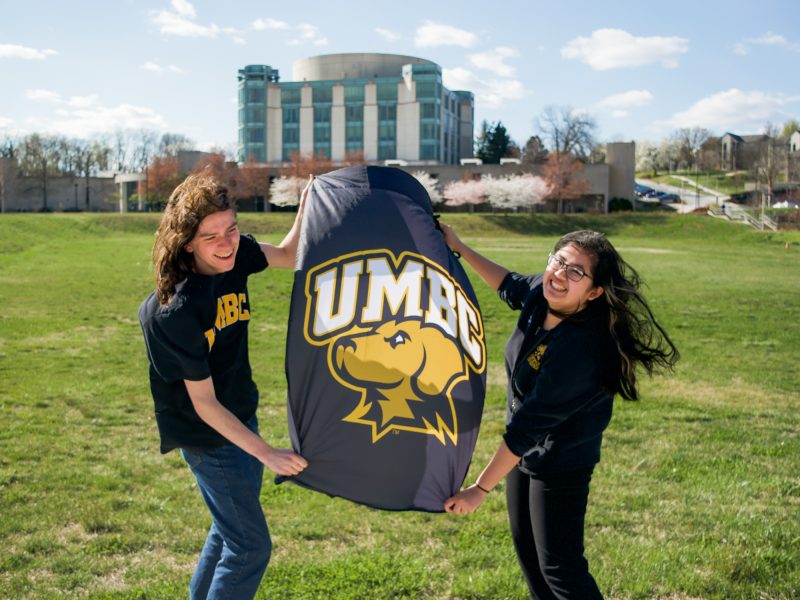 Two smiling students holding up a UMBC flag with the library in the background