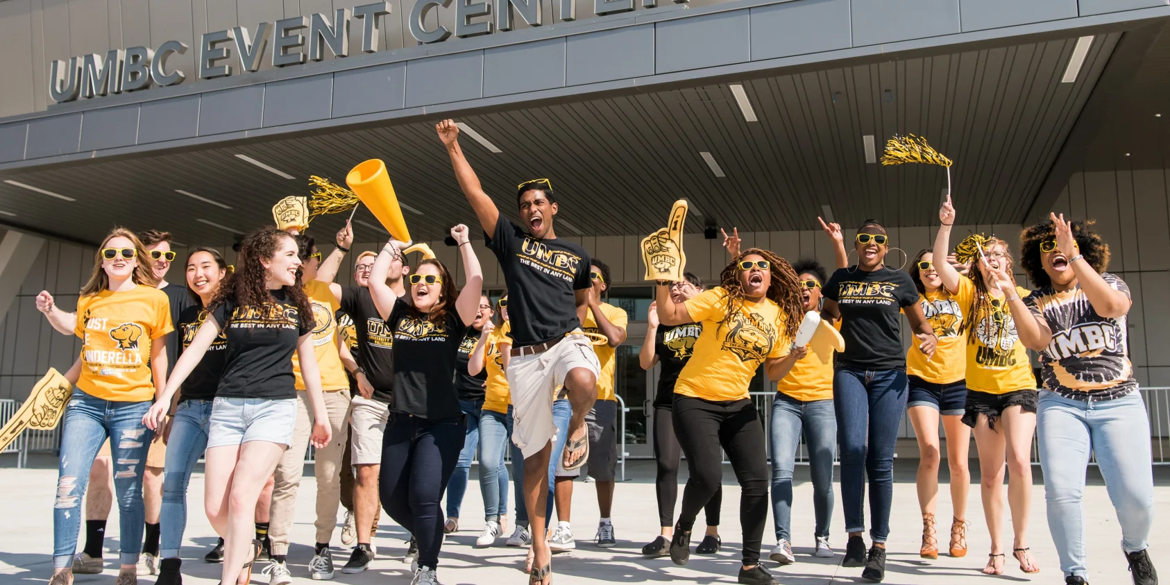 Students with pom poms and black and gold gear cheering and jumping in front of UMBC's arena
