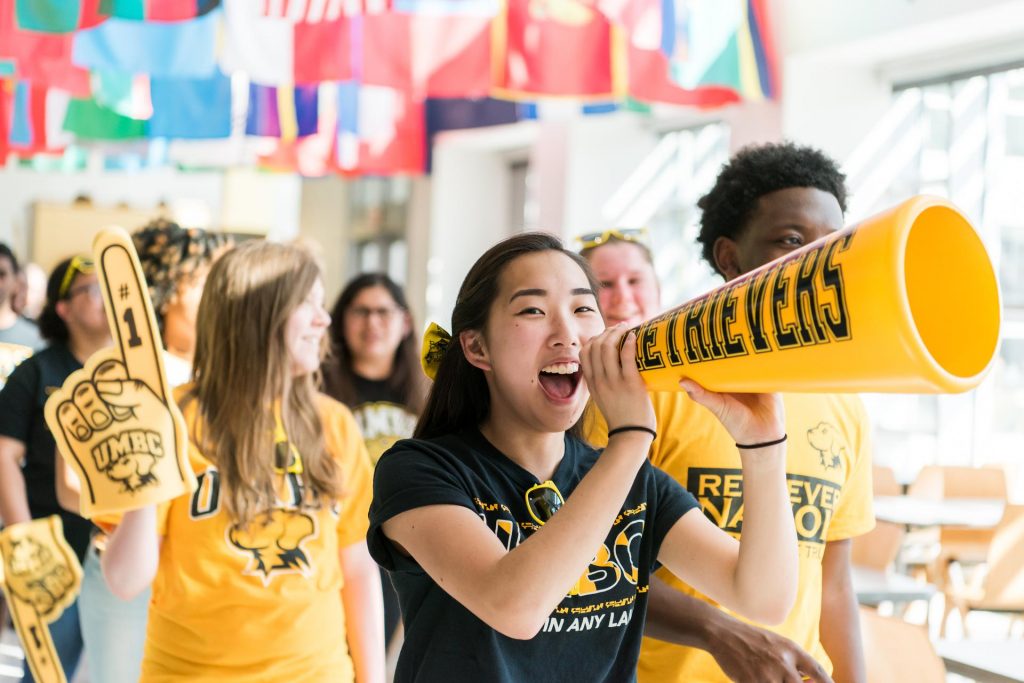 Students decked out in black and gold UMBC gear cheering and using a megaphone in the Commons