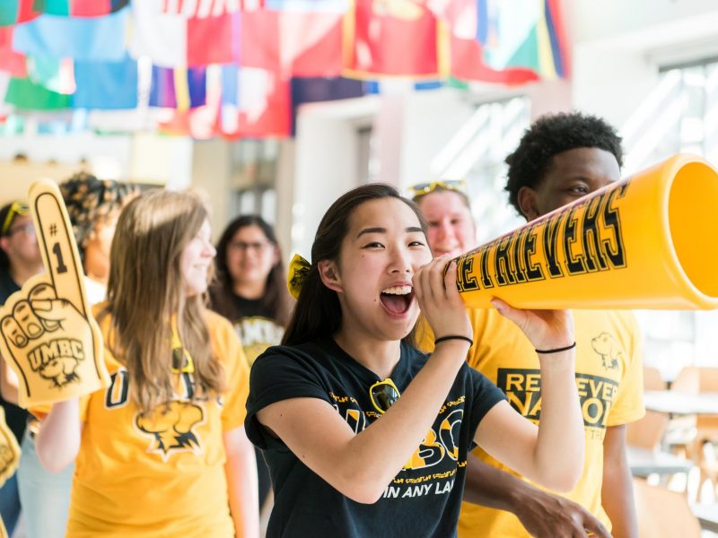 Students decked out in black and gold UMBC gear cheering and using a megaphone in the Commons