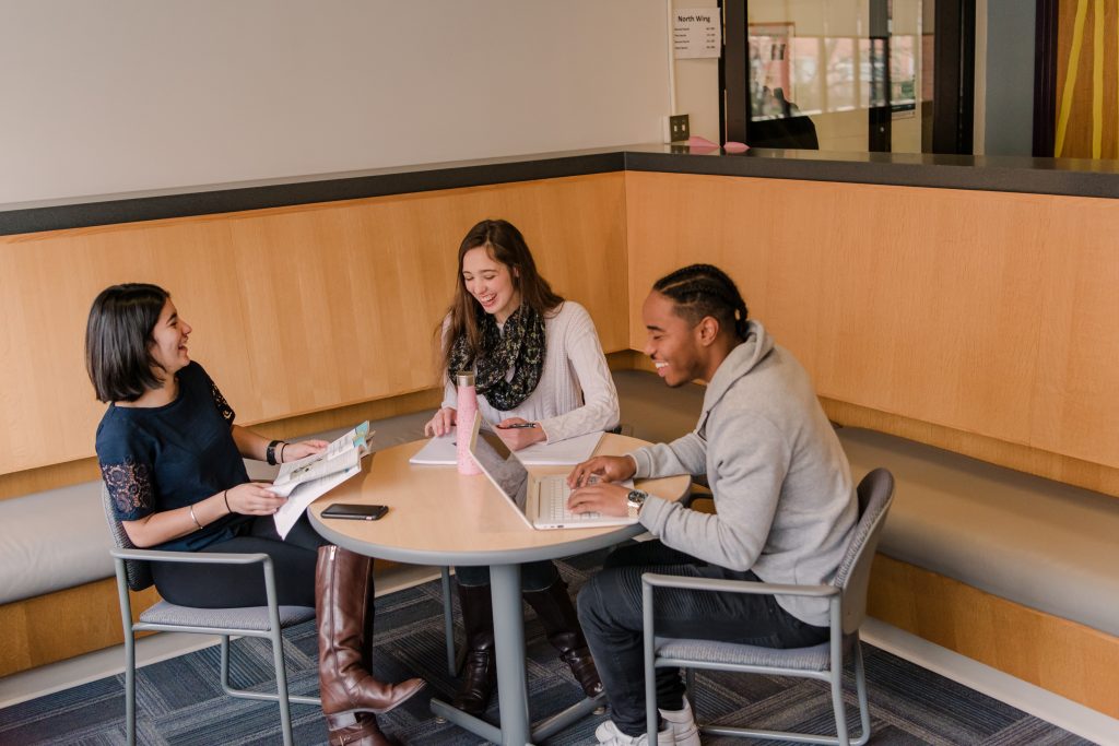 Three students sit in a residence hall lounge and laugh while doing homework