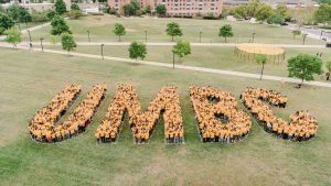A group of students stands in a grassy field forming the letters UMBC.