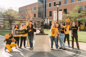 A group of students standing together with an outdoor sculpture, all wearing UMBC spirit wear in black and gold