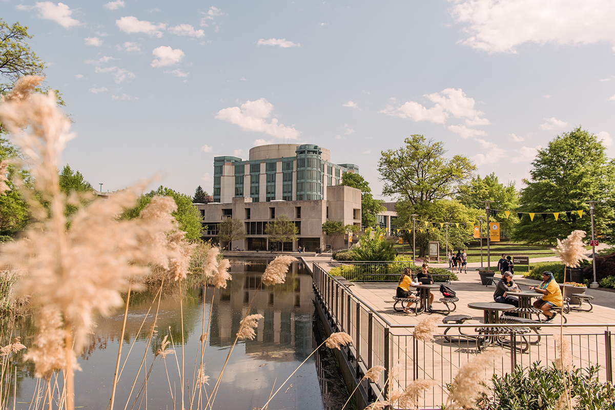 UMBC's beautiful campus, featuring the AOK Library and reflective pond. Students are sitting outside at tables.