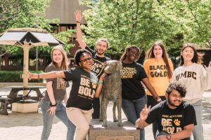 A diverse group of students wearing UMBC black and gold shirts, posing with the True Grit statue, UMBC's Chesapeake Bay Retriever mascot.